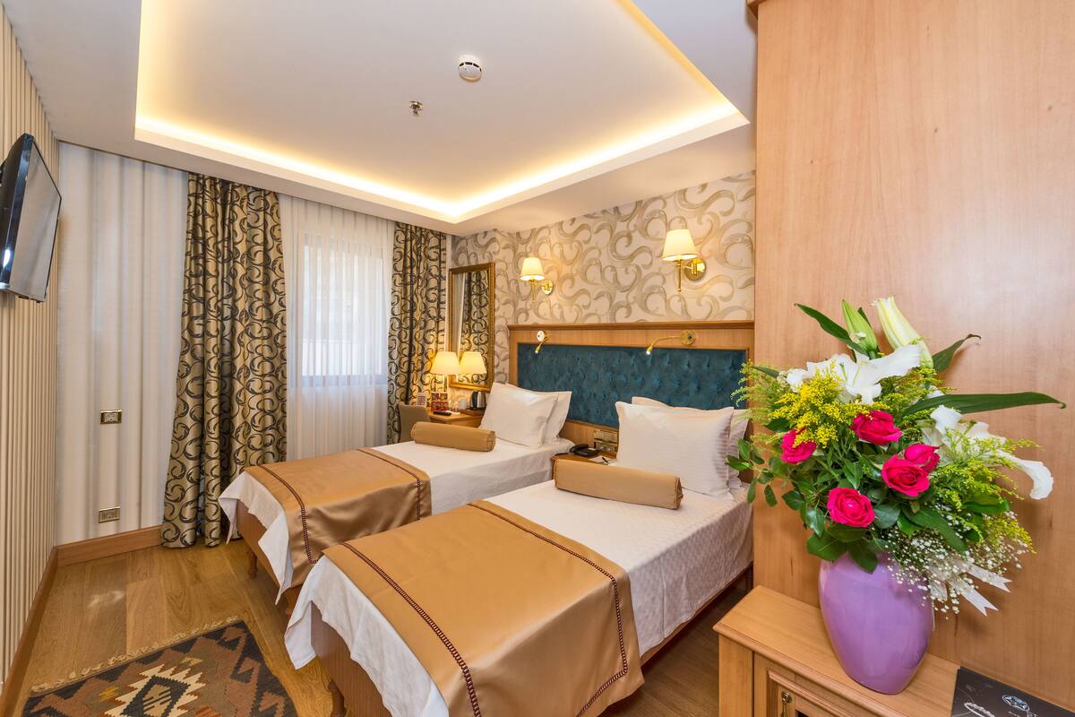 A hotel room with two beds, a wooden headboard, a nightstand, a lamp, and a vase of flowers.
