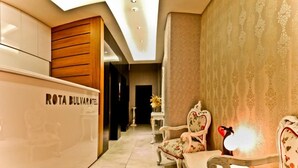 A hotel lobby with a reception desk labeled "ROTA BULVAROTEL," a floral armchair, a small table with a lamp, and a decorative wall. - Rota Bulvar Hotel (Ankara)