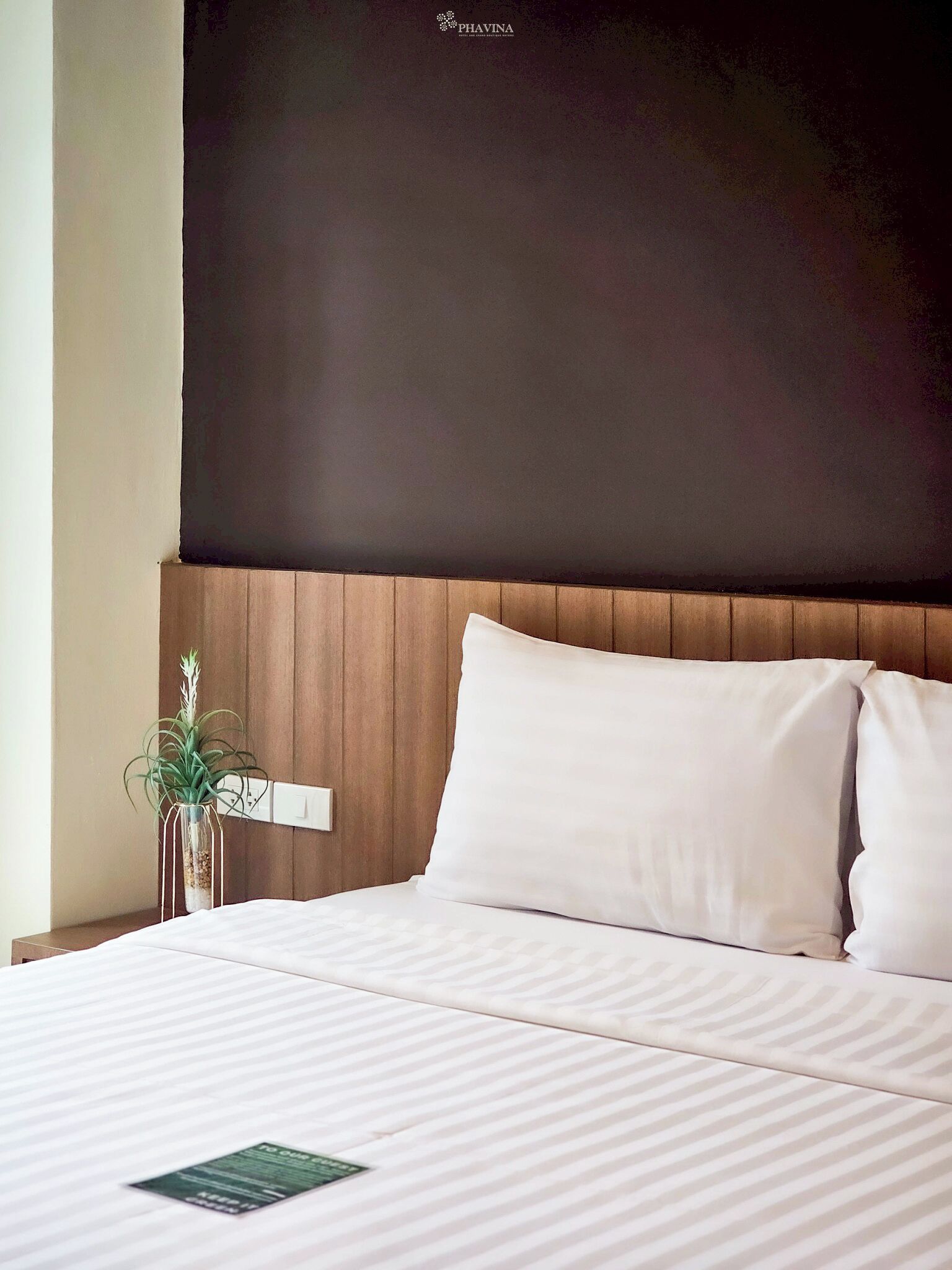 Ein Hotelzimmer mit Bett, weißer Bettwäsche, einem Holzkopfteil, einer Zimmerpflanze und einem an der Wand montierten Lichtschalter.