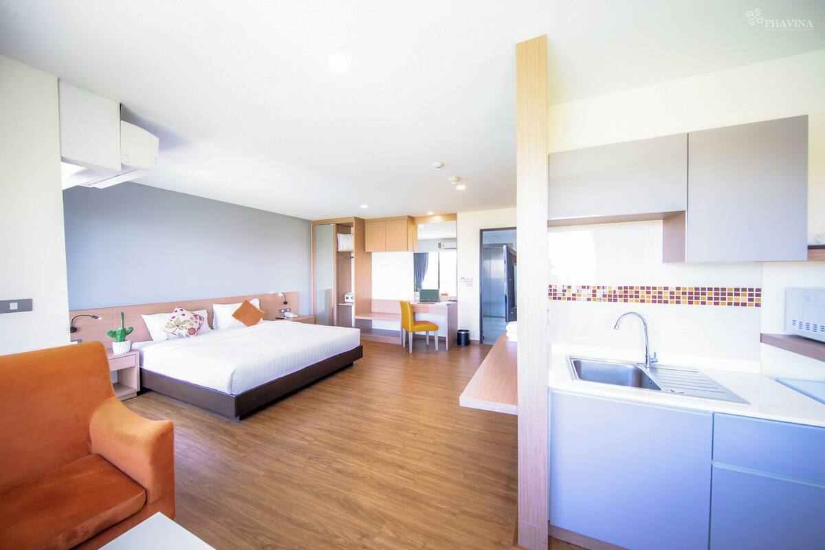 A modern hotel room with a bed, a desk, a chair, and a kitchenette.