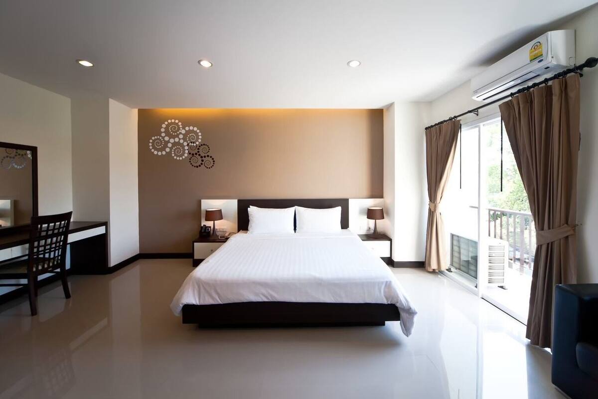 A hotel room with a large bed, bedside tables with lamps, a desk with a chair, a decorative wall piece, and a large window with curtains.