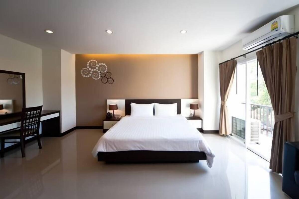 A hotel room with a large bed, bedside tables, a desk with a chair, a wall-mounted artwork, and a balcony with curtains.