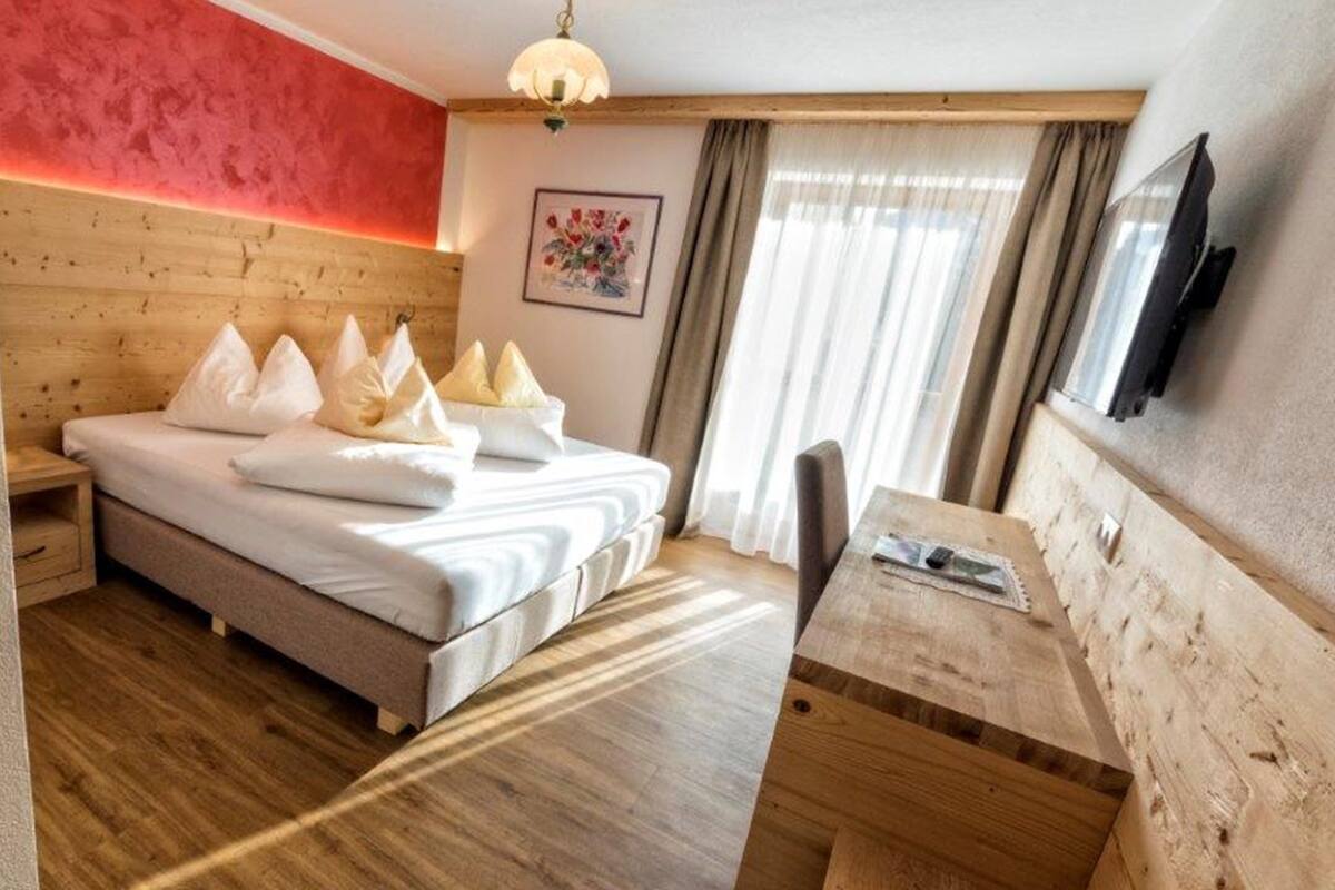 Una habitación de hotel con cabecera de madera, cama con ropa blanca, mesita de noche, escritorio y un amplio ventanal con cortinas.