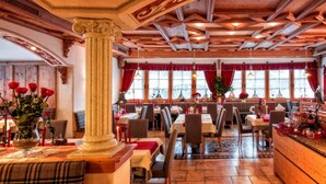 A restaurant with wooden beams, red curtains, and a central column. Tables are set with white tablecloths and red napkins. - Hotel Mirabel (Marebbe)