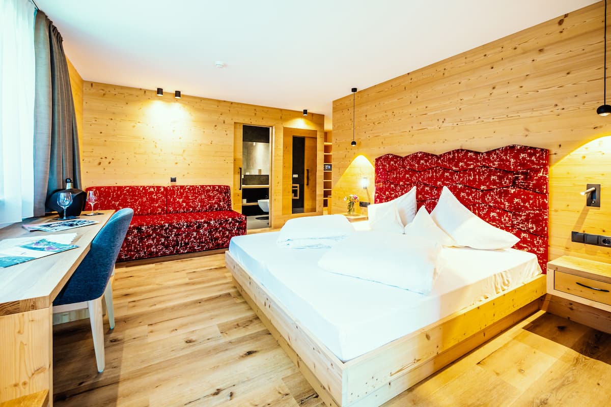 Habitación de hotel moderna con piso de madera, cama con ropa blanca, sofá tapizado rojo, escritorio con silla y cabecera de madera con respaldo rojo estampado.