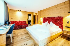 A modern hotel room with a wooden floor, a bed with white linens, a red upholstered sofa, a desk with a chair, and a wooden headboard with a red patterned backrest. - Hotel Mirabel (Marebbe)