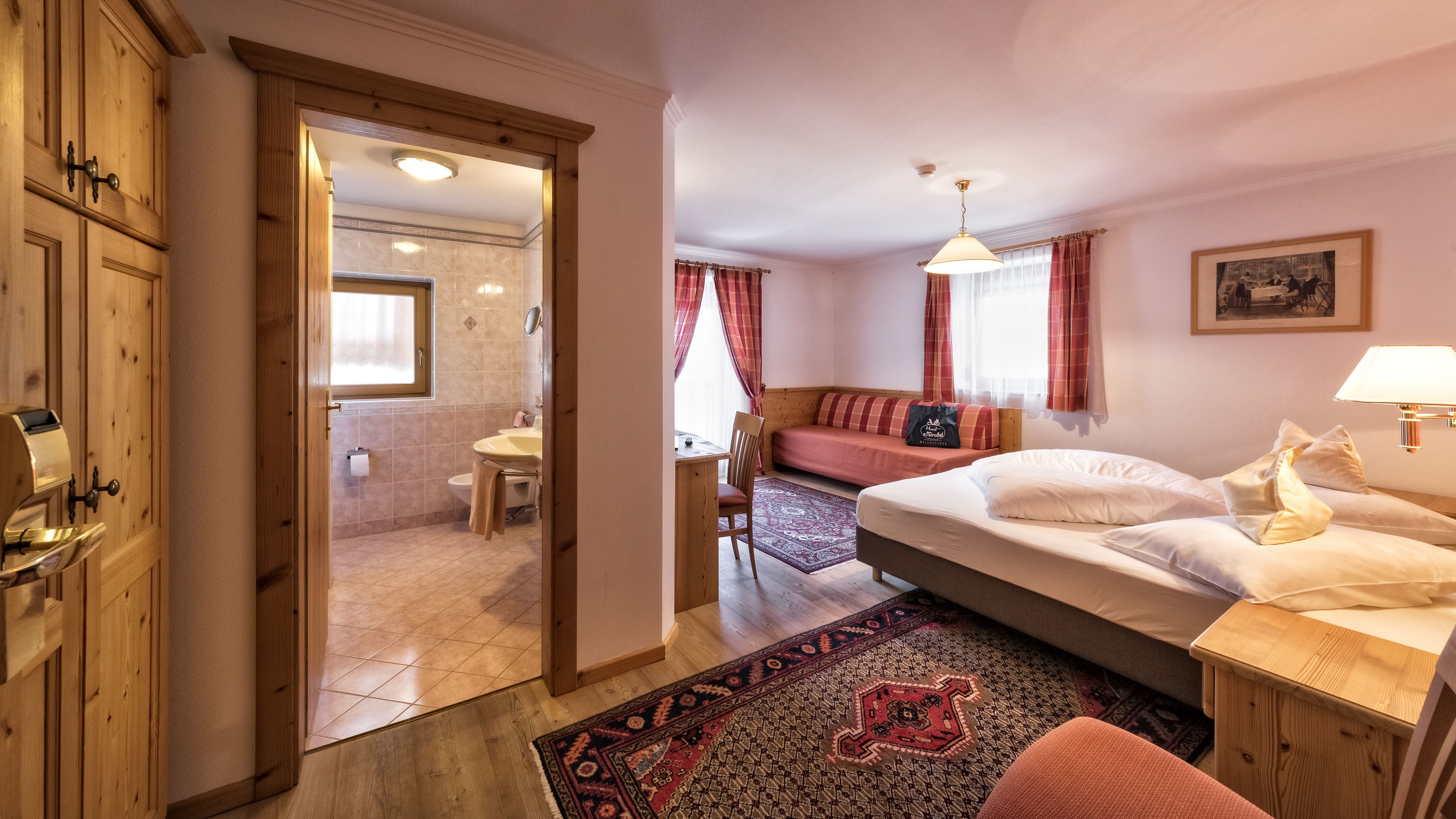 A hotel room with a wooden wardrobe, a bed with white linens, a small table with a chair, a bathroom with a toilet and sink, and a window with curtains.