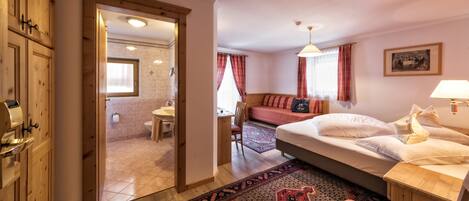 A hotel room with a wooden wardrobe, a bed with white linens, a small table with a chair, a bathroom with a toilet and sink, and a window with curtains.