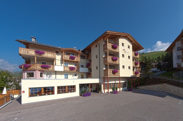 A multi-story hotel with balconies adorned with purple flowers, a clear sky, and a hilly landscape in the background. - Hotel Mirabel (Marebbe)
