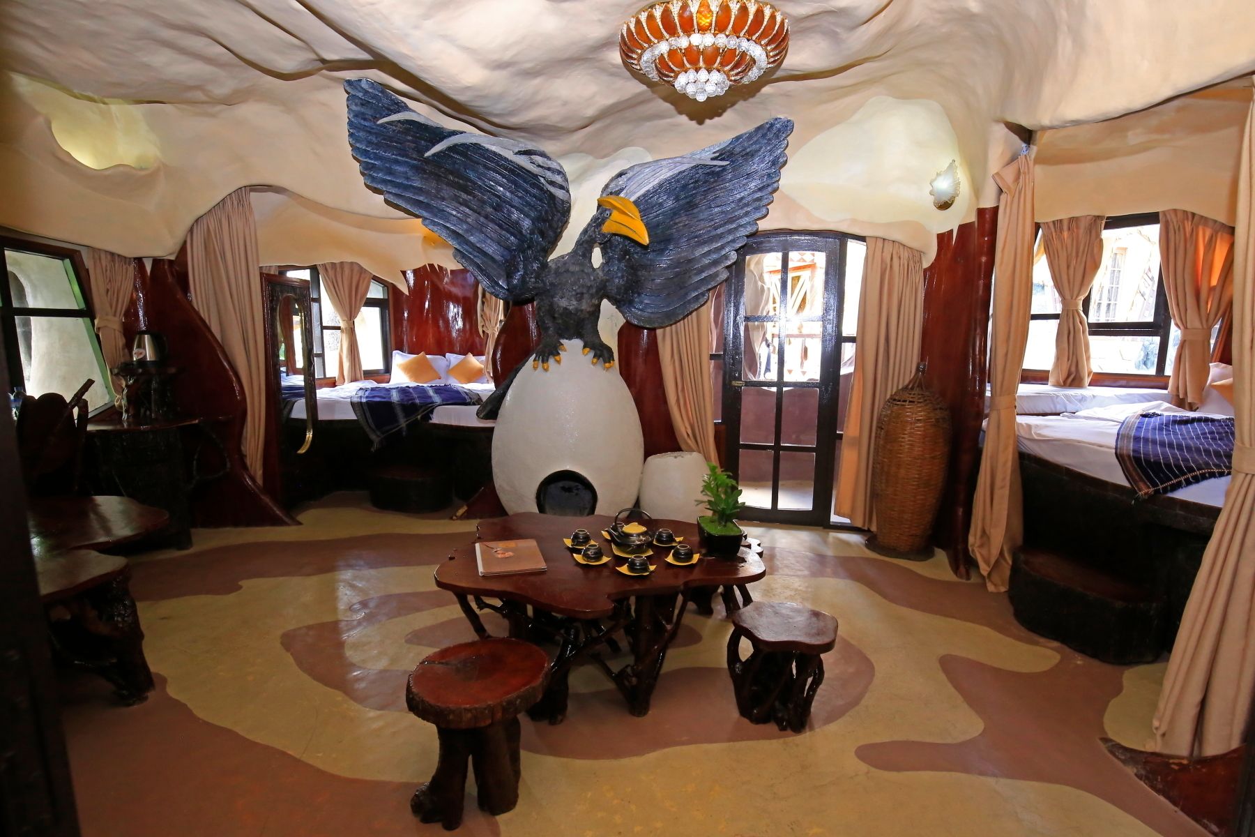 A room with a large bird sculpture, a dining table with chairs, and a bed with curtains.