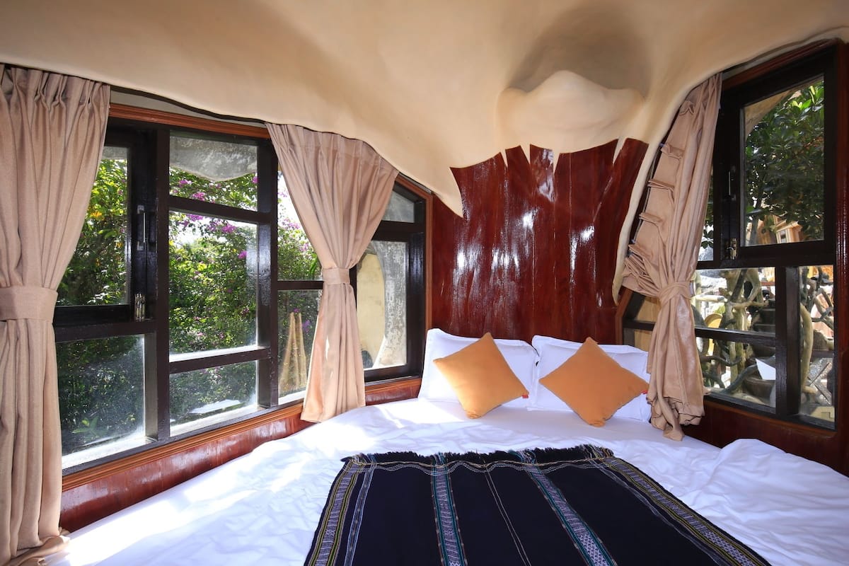 A bedroom with a large bed, beige curtains, and windows with a view of greenery.