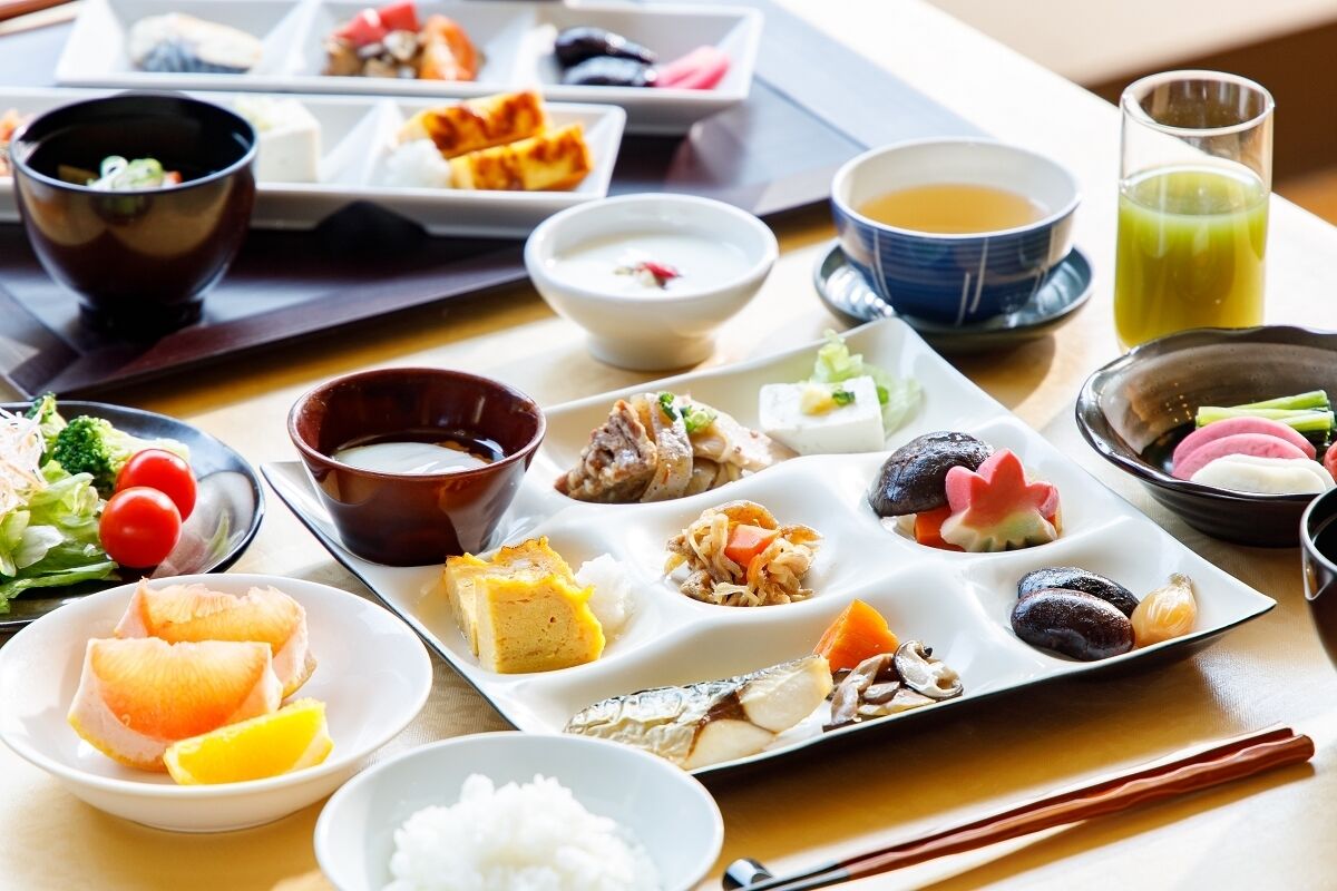 A variety of Japanese dishes on a tray, including sushi, sashimi, and pickled vegetables, accompanied by bowls of soup and a glass of green tea.
