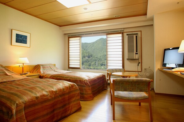 Habitación de hotel con dos camas, un escritorio con silla, televisión y ventana con vista a una montaña boscosa.