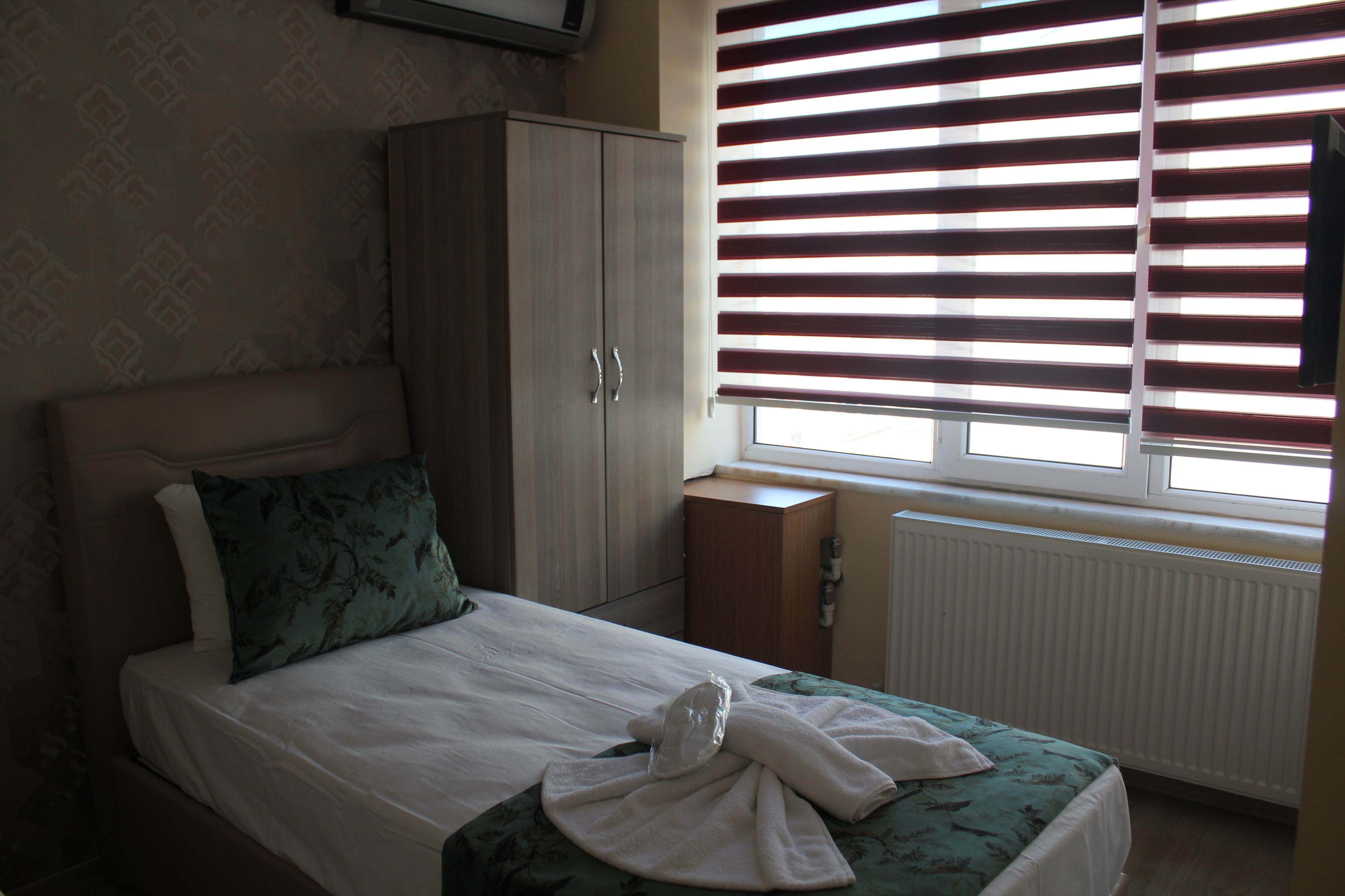 Ein Schlafzimmer mit Bett, Schrank, Heizkörper und einem Fenster mit Jalousien.