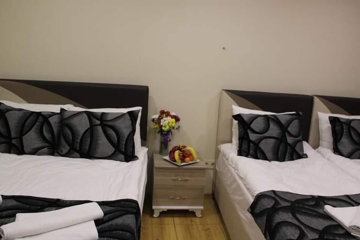 A hotel room with two beds, each with black and white patterned bedding, a small wooden nightstand with a vase of flowers and fruit, and a plain wall.