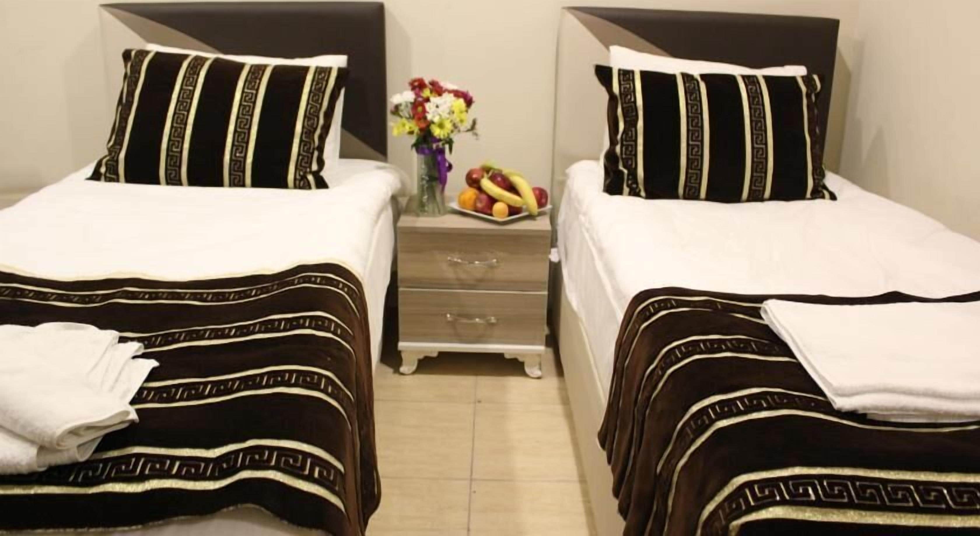 Habitación de hotel con dos camas, cada una con colcha a rayas negras y blancas, una mesita de noche con un florero y fruta, y un mueble cajonero de madera.