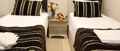 Habitación de hotel con dos camas, cada una con colcha a rayas negras y blancas, una mesita de noche con un florero y fruta, y un mueble cajonero de madera.