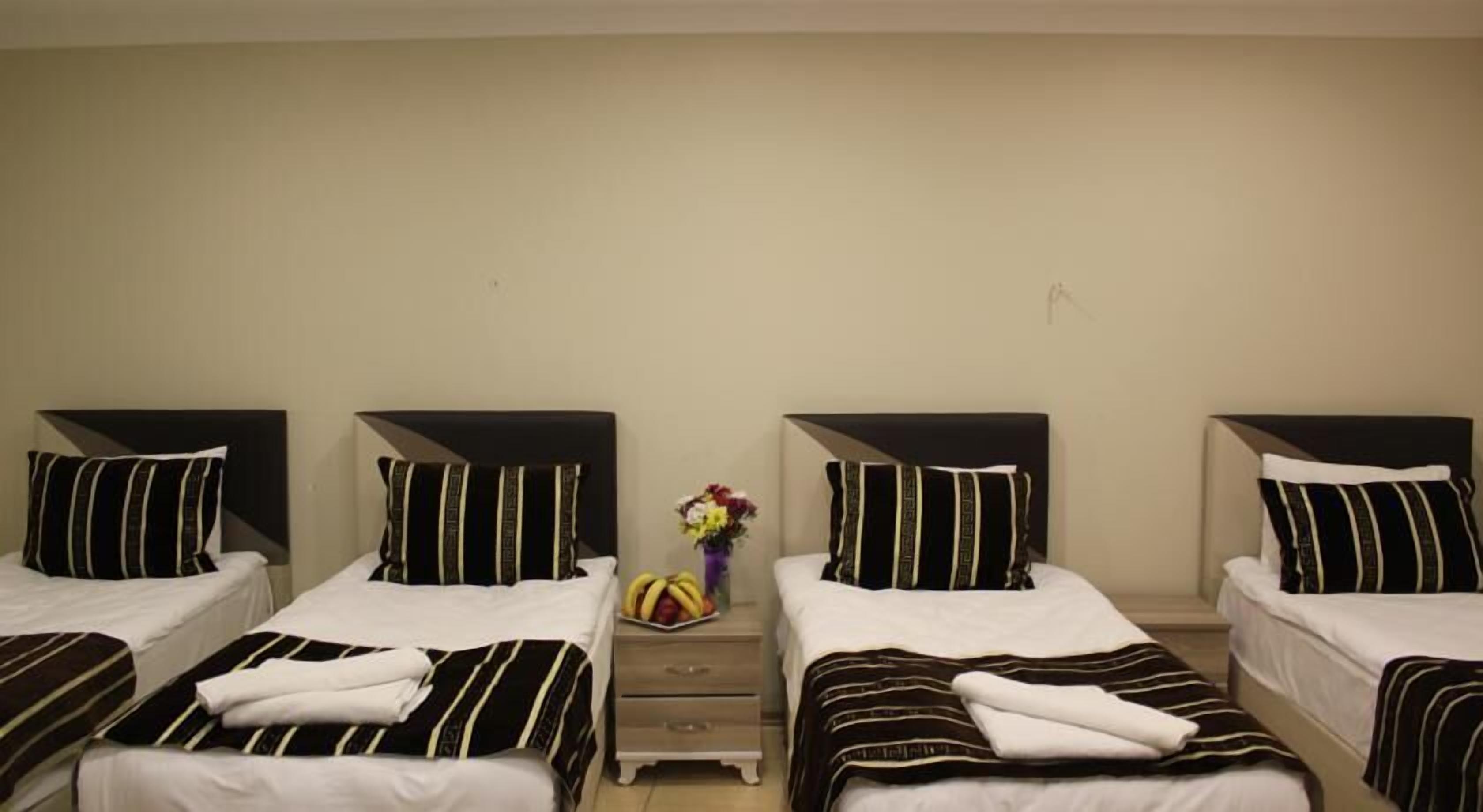 A hotel room with four beds arranged in a row, each with black and white striped bedding and matching headboards. There is a small wooden nightstand between the beds with a vase of flowers and a fruit bowl.