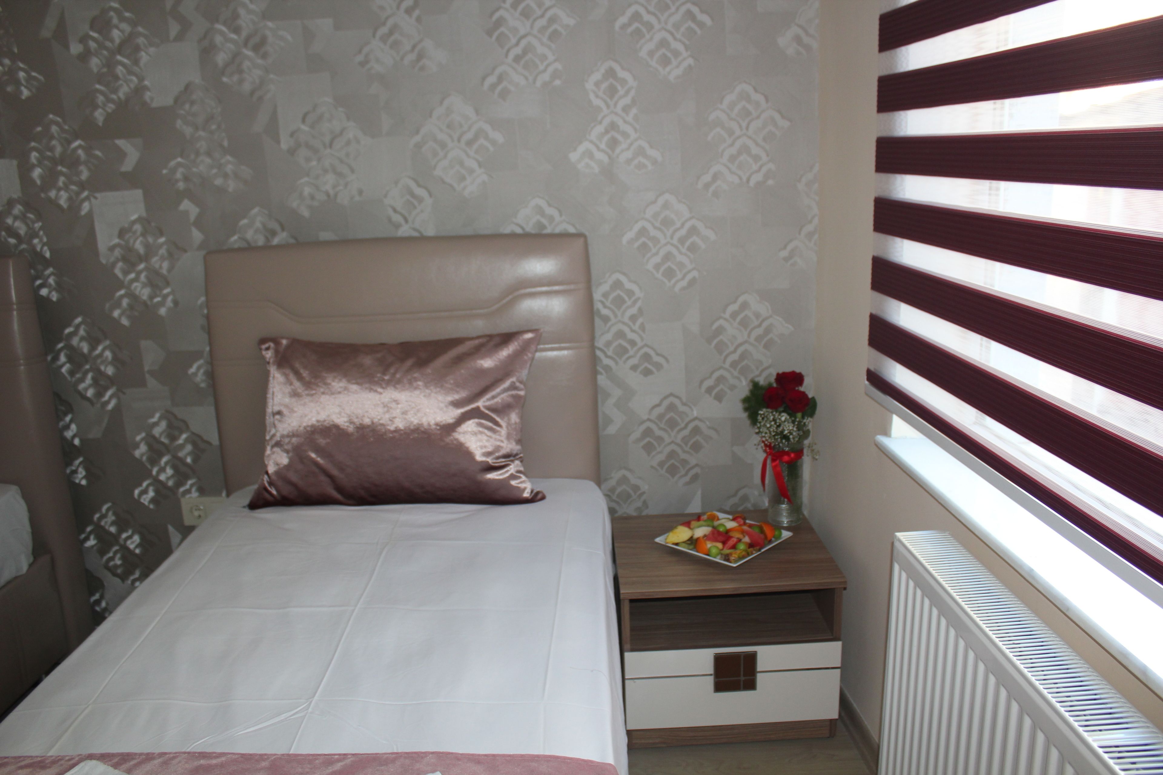A neatly arranged bedroom with a bed, a bedside table with a vase of flowers, and a window with blinds.