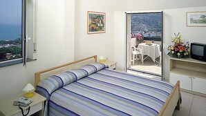 Een slaapkamer met een bed, een nachtkastje met een lamp, een stoel, een klein tafeltje met een bloevas en een balkon met een tafel en stoelen.