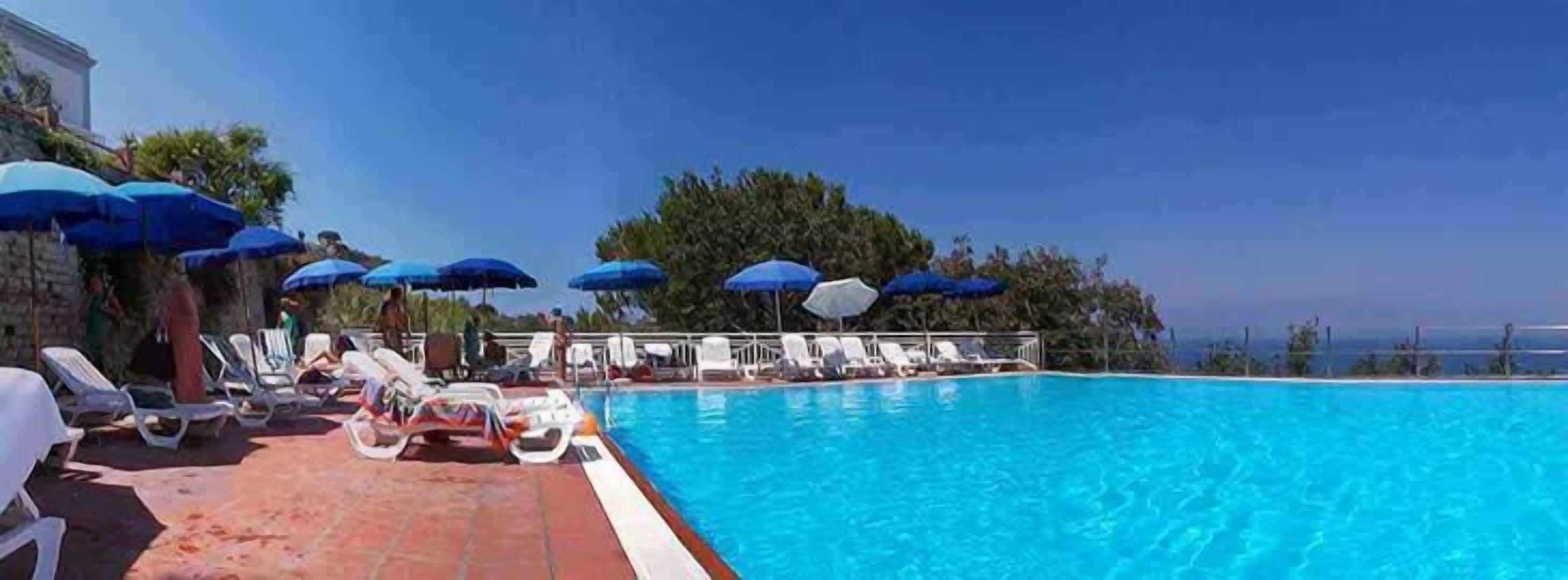 A poolside area with lounge chairs and umbrellas, a clear blue pool, and a view of the sea.