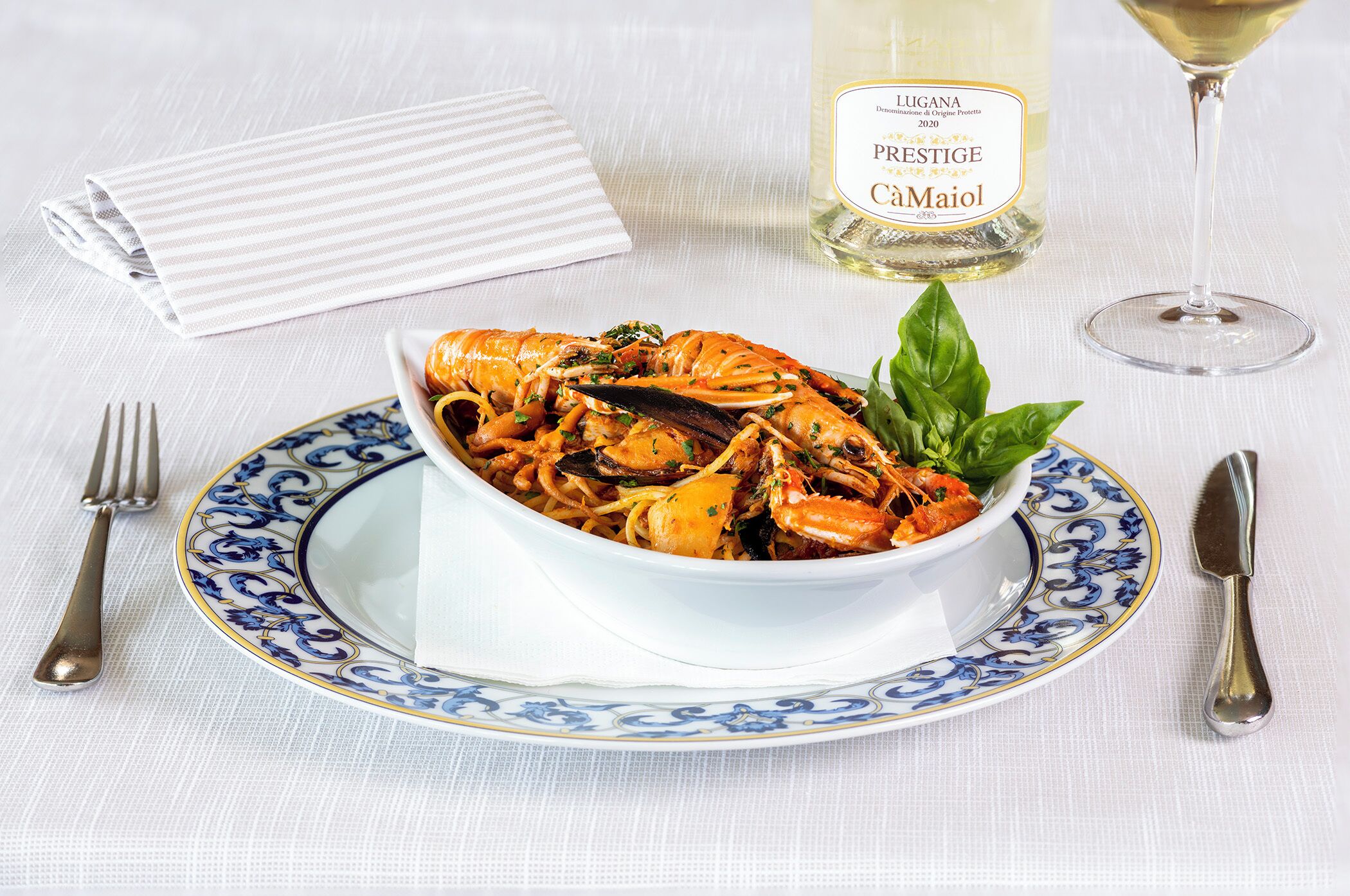 A bowl of seafood pasta on a decorative plate, a bottle of wine, and a glass of wine on a tablecloth.