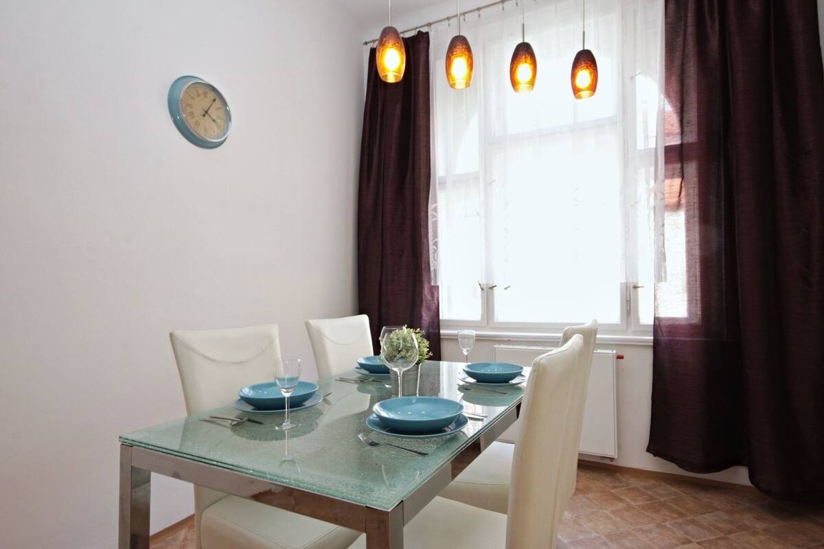Uma área de refeições com mesa de vidro, cadeiras brancas e pratos azuis. Há luminárias pendentes e uma janela com cortinas.
