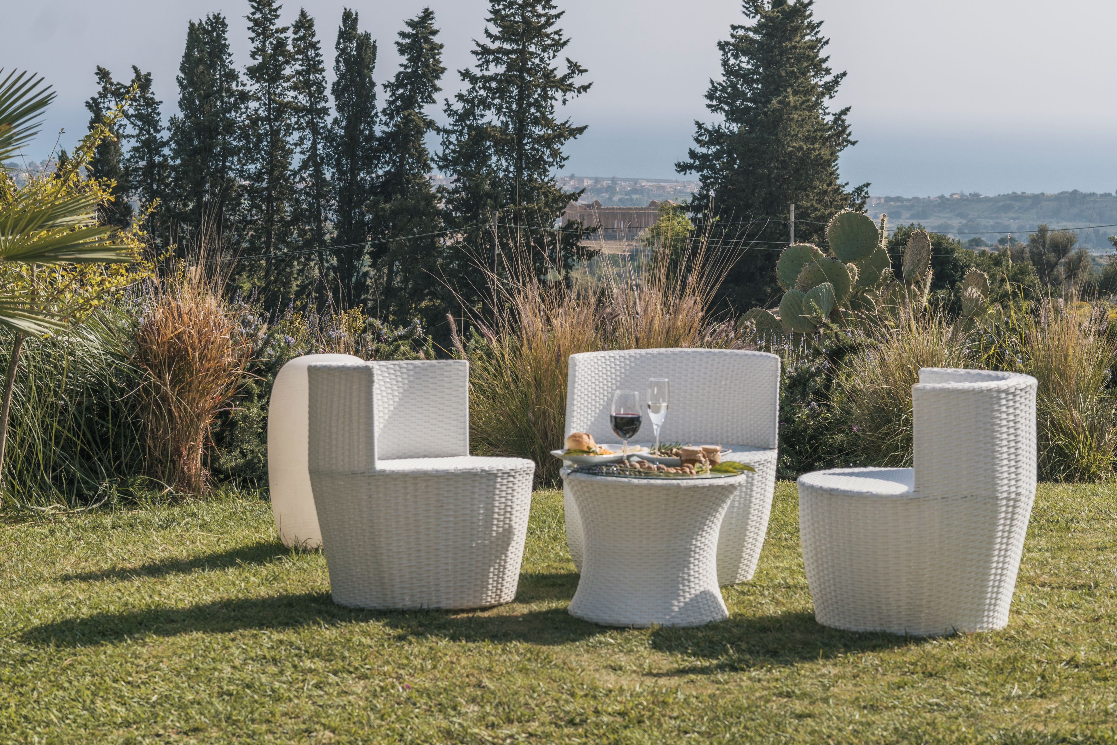 An outdoor seating area with three white wicker chairs and a round table, set on a grassy lawn with a view of trees and a distant landscape.
