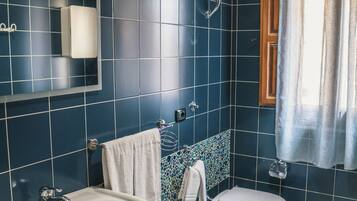 A bathroom with dark blue tiles, a white sink, two toilets, and a small wicker stool.