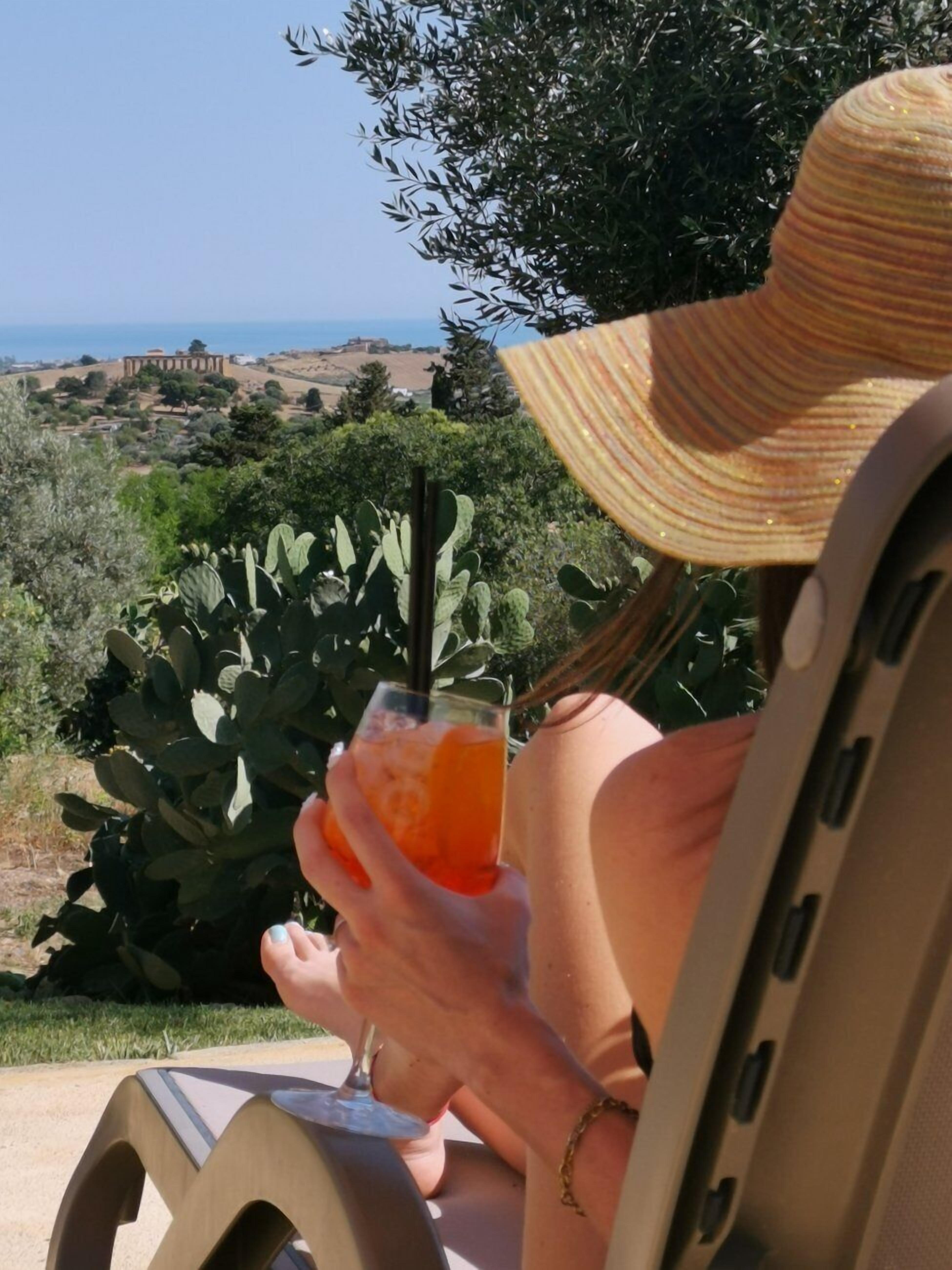 A person relaxing on a lounge chair with a drink, overlooking a landscape with cacti and a distant view of a building.