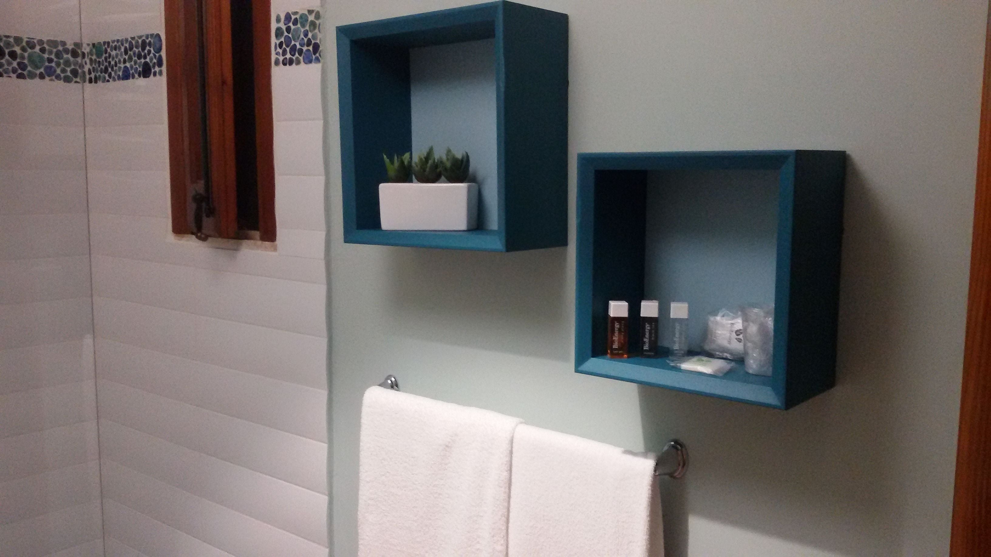 Un baño con paredes de azulejos blancos, una toalla blanca colgada en un perchero y dos estantes azules con artículos de aseo.
