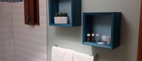 Un baño con paredes de azulejos blancos, una toalla blanca colgada en un perchero y dos estantes azules con artículos de aseo.
