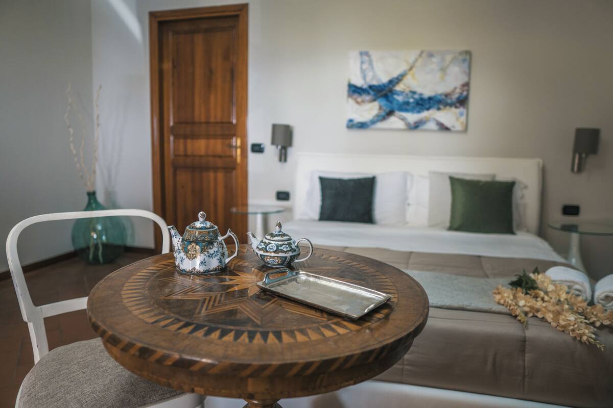 Een kamer met een ronde houten tafel, een witte stoel, een bed met groen-witte beddengoed, een schilderij aan de muur en een houten deur.