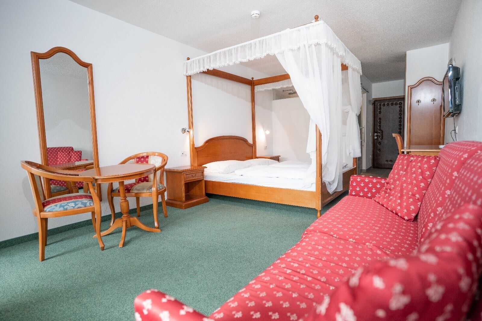 Um quarto com uma cama de dossel, uma cadeira de madeira, uma mesinha e um sofá vermelho.