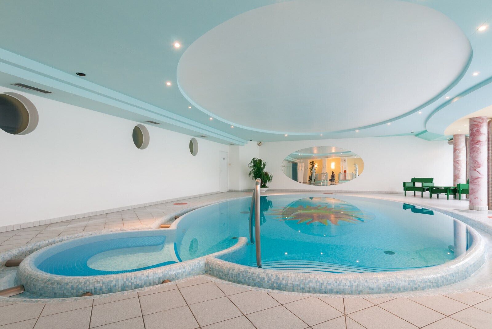 An indoor swimming pool with a tiled floor, a central water feature, and a seating area with green chairs.