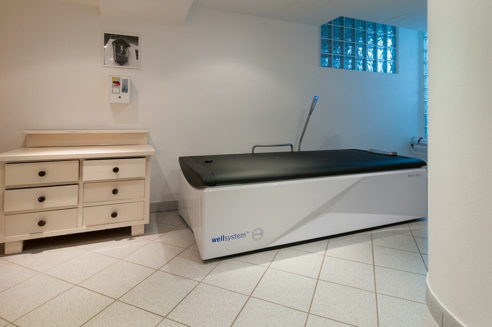 Een steriele behandelkamer met een witte onderzoeksbank, een wit kastje met lades en een raam met blauw getint glas.