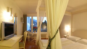 A hotel room with a bed, a desk with a television, a chair, a window with curtains, and a nightstand with a lamp. - Hotel Panoramik (Rio di Pusteria)