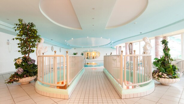 A spacious indoor swimming pool area with tiled floors, white railings, and decorative plants. - Hotel Panoramik (Rio di Pusteria)