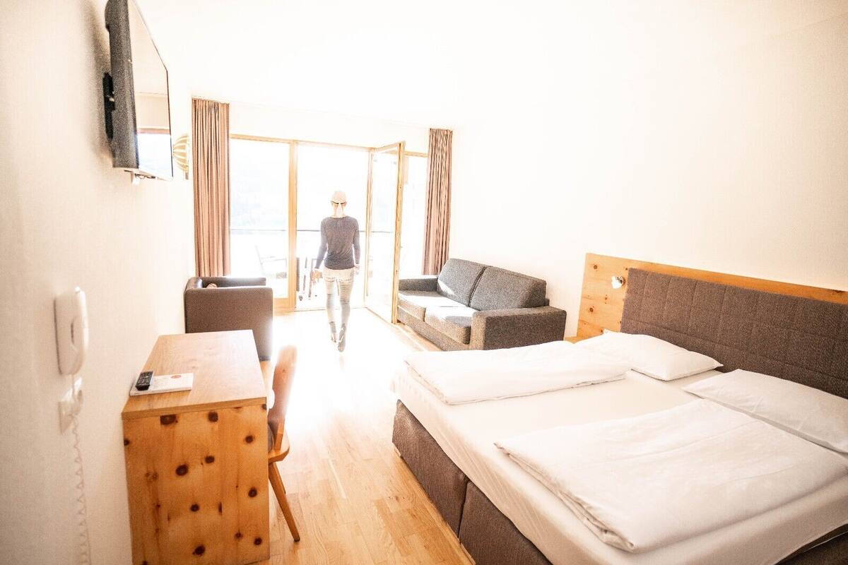 A hotel room with two beds, a desk, a chair, a sofa, and a large window with curtains.