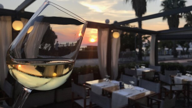 Una copa de vino blanco con vistas a un atardecer a través de un techo de cristal, con vista a un comedor con mesas y sillas.