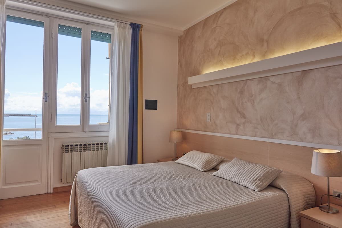 Une chambre d’hôtel avec un grand lit, une table de chevet avec une lampe, une vue sur la mer par la fenêtre et une tête de lit en bois.