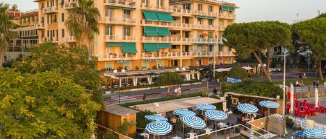 Un hotel de varios pisos con balcones, mobiliario exterior con sombrillas y vista a la calle.