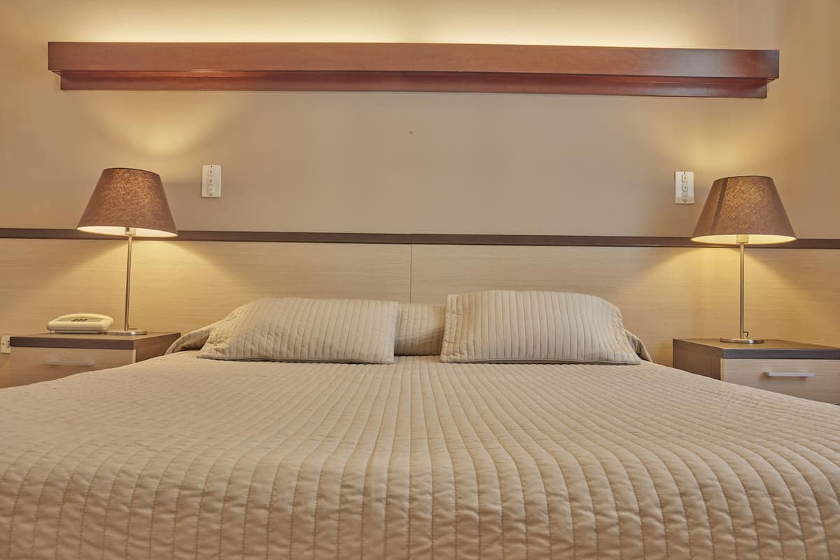 Un lit bien fait, recouvert d’une courtepointe beige, avec deux lampes de chevet et une tête de lit en bois.