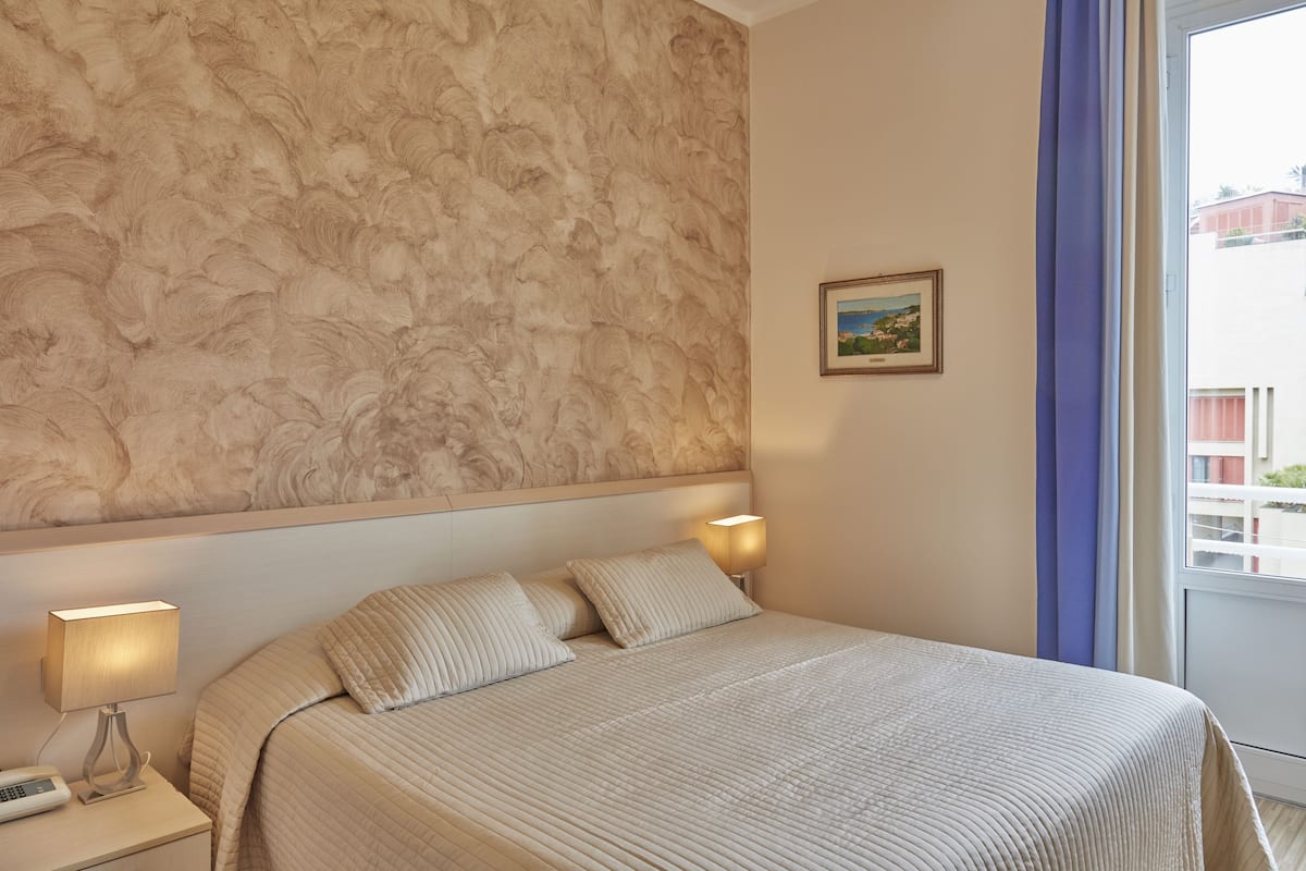 Une chambre d’hôtel avec un grand lit, deux lampes de chevet, une table de nuit, un mur orné d’un motif texturé, un tableau encadré et une fenêtre avec des rideaux.