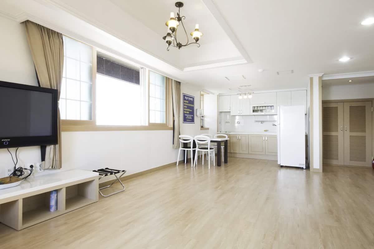 Amplia sala de estar con una televisión de pantalla plana, una mesa de comedor con sillas, una cocina al fondo y un pasillo que conduce a una puerta.