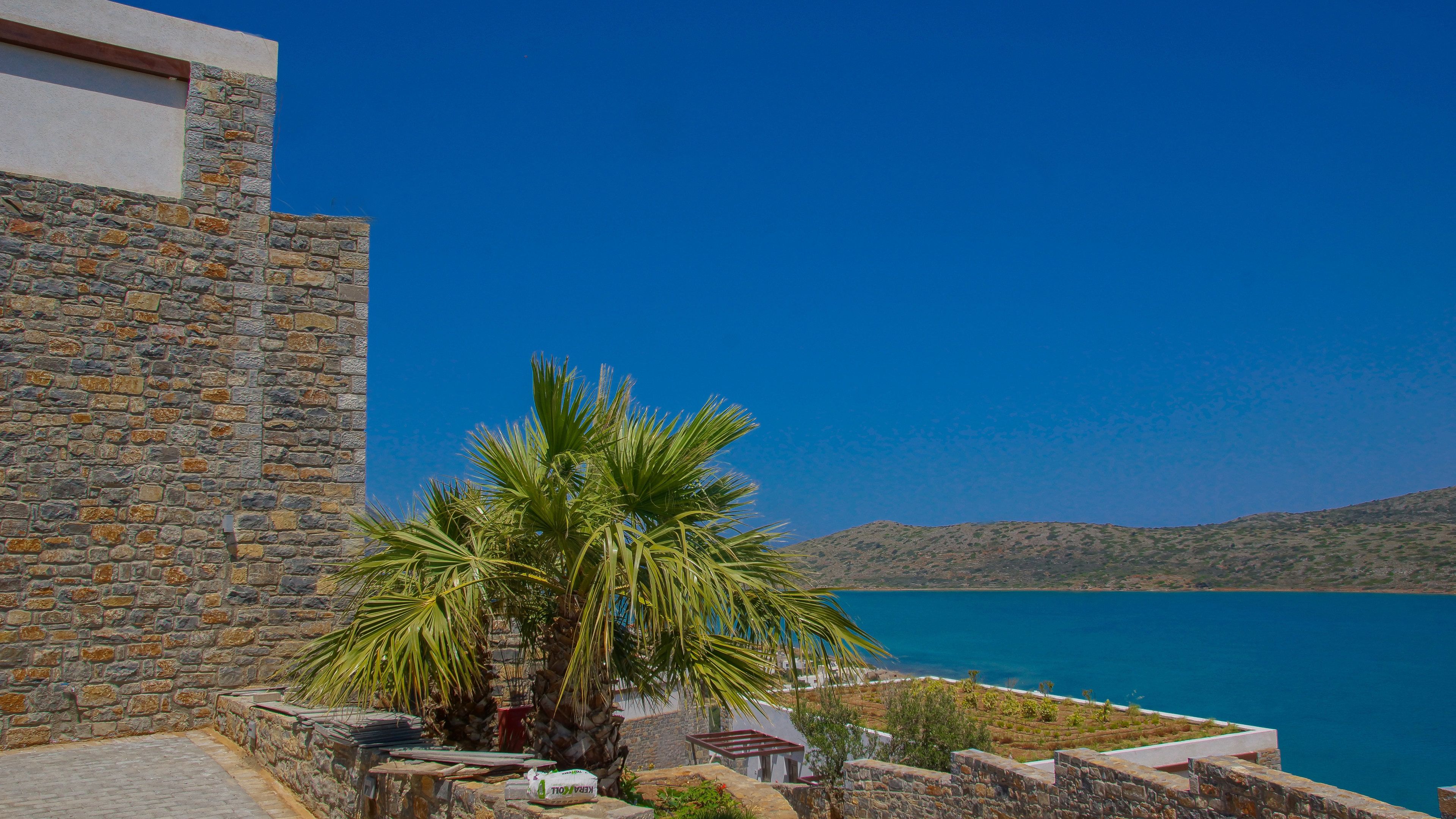 Un edificio de piedra con una palmera frente a él, con vistas al mar azul y un paisaje montañoso.