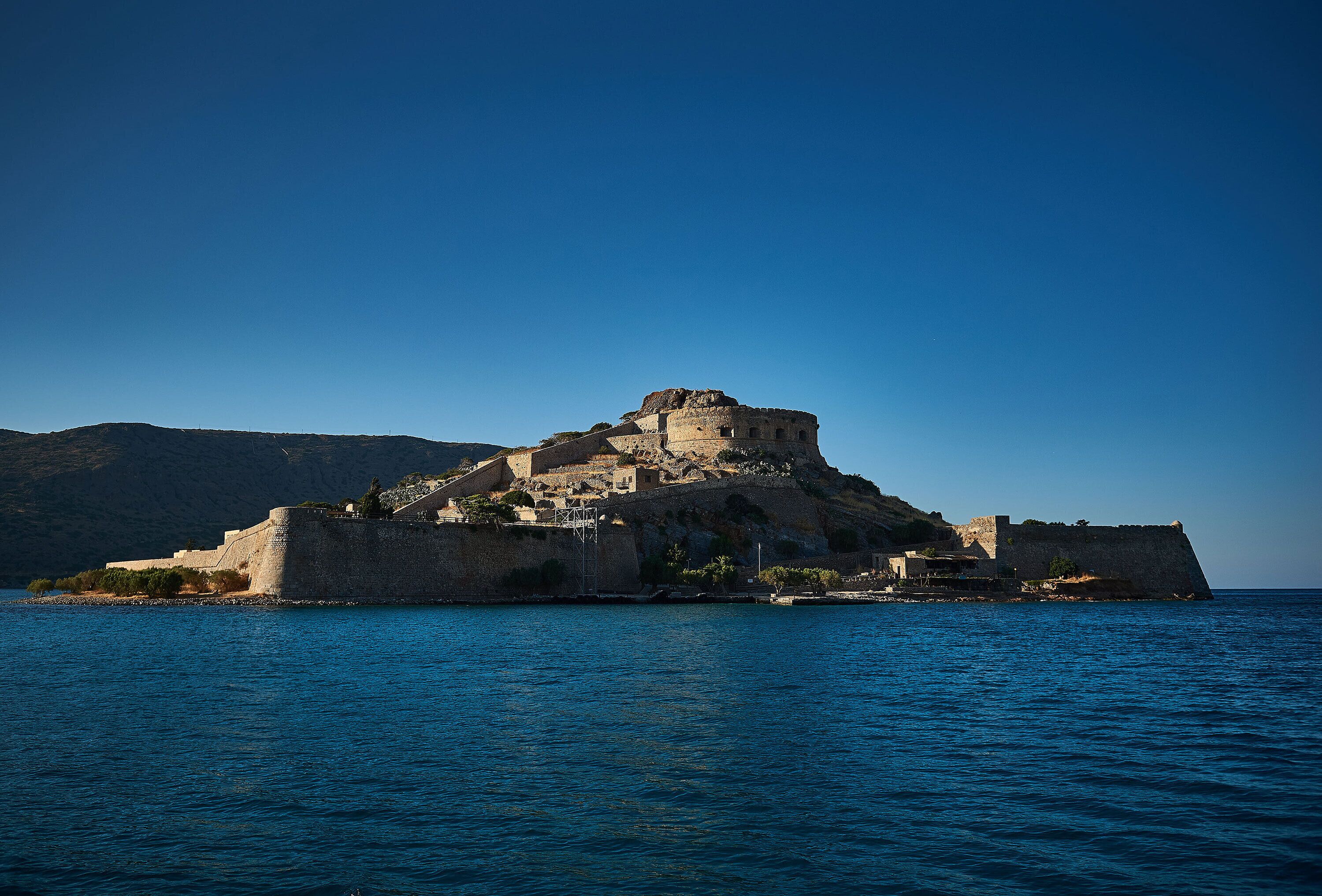 A coastal fortification with a prominent central tower, surrounded by walls and smaller structures, overlooking a calm sea.