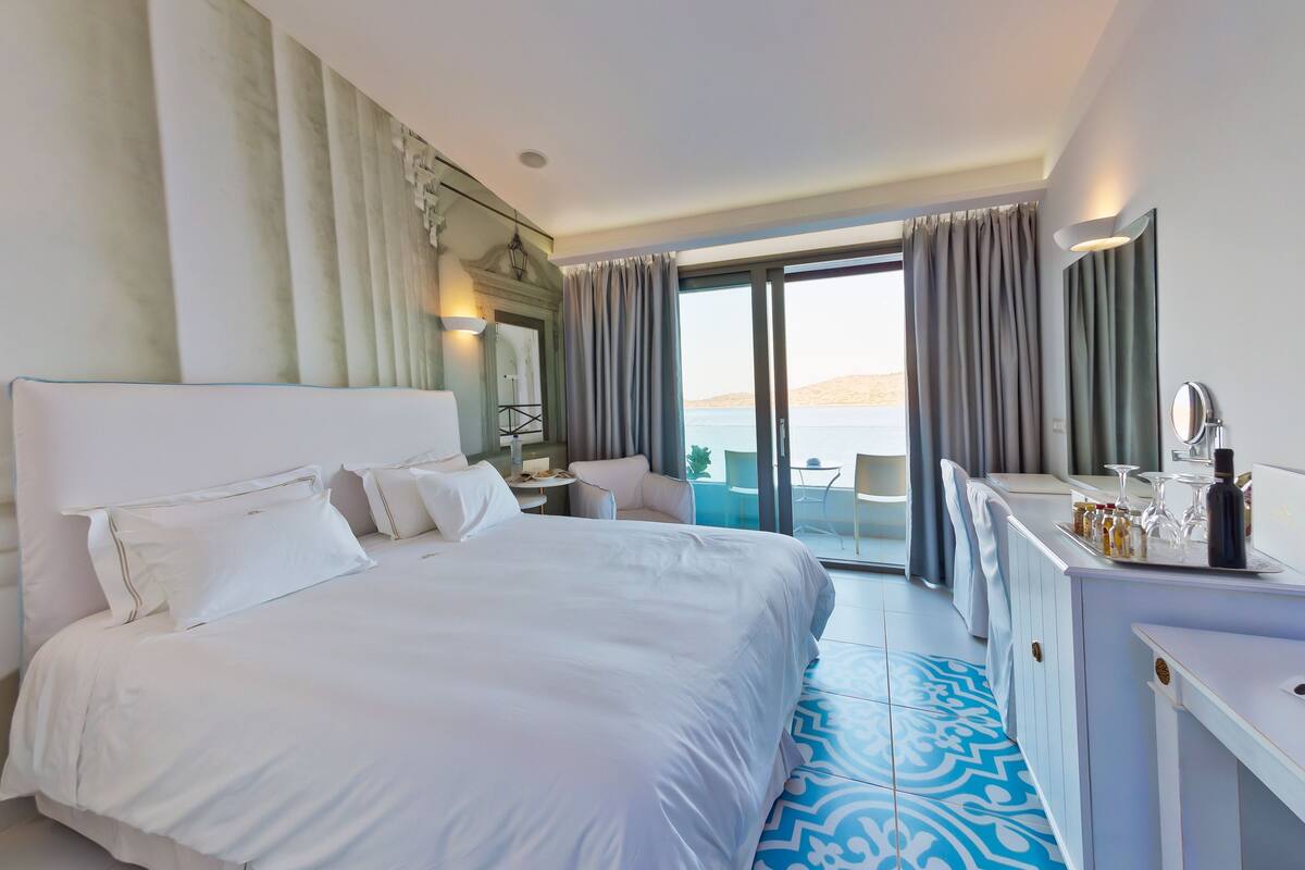 Une chambre d’hôtel avec un grand lit, un bureau avec un miroir et une vue sur la mer, accessible par une porte-fenêtre coulissante.