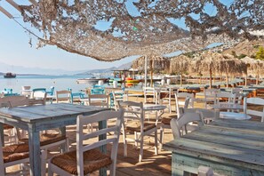 An outdoor seating area with tables and chairs, a view of the sea, and thatched umbrellas.
