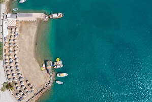 Vista aérea de un hotel frente al mar, con filas de tumbonas, un muelle y varios barcos.